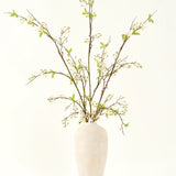 Een hoge, crèmekleurige vaas van keramiek bevat het Zijden kunstbloemen boeket Verbena crème met lange takken, kleine groene blaadjes en delicate witte bloesems, prachtig gerangschikt tegen een effen lichte achtergrond.