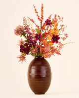 Zijden kunstbloemenboeket Amber Bloom, bestaande uit oranje, paarse en rode zijden bloemen in een bruine, geribbelde vaas.