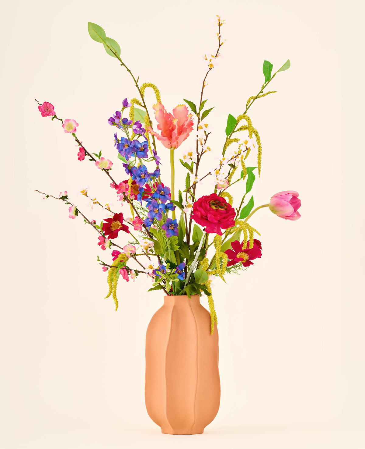 Een April Joy-boeket van kleurrijke zijden bloemen, waaronder roze tulpen en felblauwe takjes, geschikt in een perzikkleurige vaas.