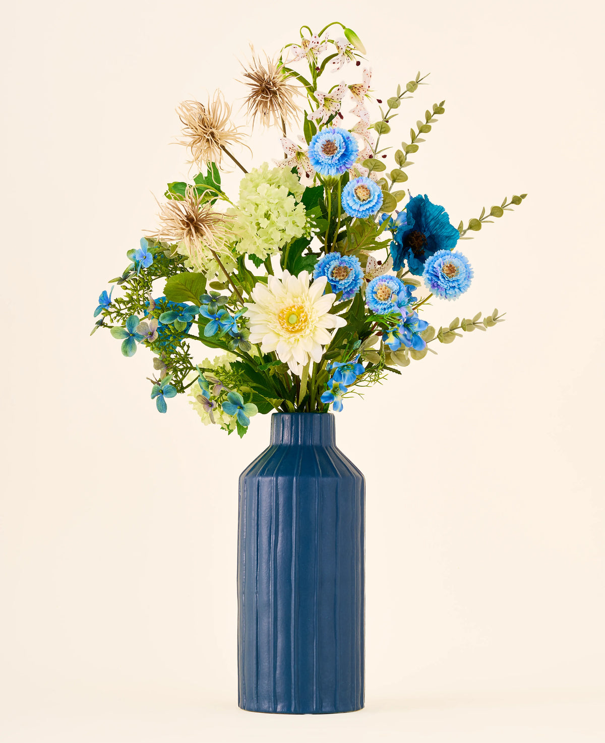 Een boeket van blauwe, witte en groene Zijden kunstbloemen, het boeket Blue Whisper, in een hoge, geribbelde blauwe vaas.