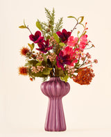 Zijden kunstbloemenboeket Cinnamon Ruby in tinten rood, roze en groen, gepresenteerd in een geribbelde, paarse vaas.