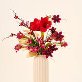 Zijden kunstbloemenboeket Rosy Noël met grote rode amaryllissen, donkerpaarse bloemen en gouden bladeren in een geribbelde, crèmekleurige vaas.