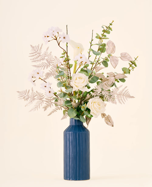 Zijden kunstbloemenboeket Silent Night, bestaande uit witte kunstrozen, calla’s en metallic bladeren in een geribbelde blauwe vaas.