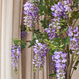 Voor de beige gordijnen hangt een kunstplant Blauweregen van 150 cm met groene bladeren.