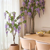 Twee Kunstplant Blauweregen van 150 cm, met hun overhangende paarse bloemen, staan naast een houten eettafel met lichte stoelen. Zacht natuurlijk licht valt door de doorschijnende gordijnen achter de planten naar binnen.