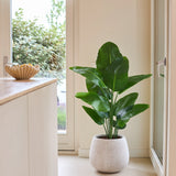 Een Kunstplant Strelitzia Real Touch Deluxe van 120 cm in weelderig groen staat op een lichte vloer naast een glazen deur in een lichte, moderne kamer met neutrale muren en een houten toonbank.
