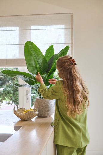 Een vrouw met lang golvend haar, gekleed in een groen pak, verzorgt een Kunstplant Strelitzia Real Touch Deluxe van 90 cm op een toonbank naast een schaal met citroenen in een lichte kamer met een raam.