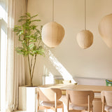 Een lichte, minimalistische eethoek is ingericht met stoelen van licht hout, een tafel, drie papieren hanglampen, de Kunstplant Mahonia van 210 cm in een pot en gordijnen die van de vloer tot aan het plafond reiken bij een groot raam dat zonlicht binnenlaat.
