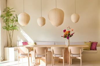 Een moderne eetkamer met stoelen van licht hout, een houten tafel, een witte zitbank, hangende papieren lantaarns, een vaas met kleurrijke bloemen en de Kunstplant Mahonia van 210 cm in een pot, die in de hoek tegen een crèmekleurige muur staat.