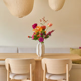 Een minimalistische eethoek met een tafel en stoelen van licht hout, een beige bank, twee grote papieren lantaarns en een witte vaas met het boeket ‘Colourful Svenski’ van Zijden kunstbloemen als levendig middelpunt. De ruimte is ingericht in zachte, neutrale tinten, wat zorgt voor een gezellige en uitnodigende sfeer.