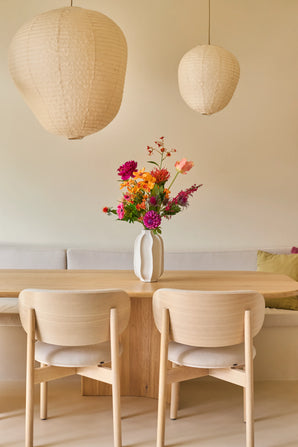 Een minimalistische eethoek met een tafel en stoelen van licht hout, een beige bank, twee grote papieren lantaarns en een witte vaas met het boeket ‘Colourful Svenski’ van Zijden kunstbloemen als levendig middelpunt. De ruimte is ingericht in zachte, neutrale tinten, wat zorgt voor een gezellige en uitnodigende sfeer.