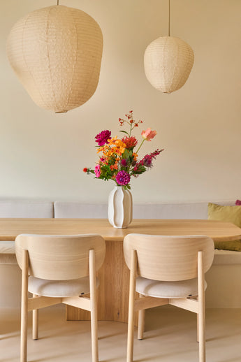 Een minimalistische eethoek met een tafel en stoelen van licht hout, een beige bank, twee grote papieren lantaarns en een witte vaas met het boeket ‘Colourful Svenski’ van Zijden kunstbloemen als levendig middelpunt. De ruimte is ingericht in zachte, neutrale tinten, wat zorgt voor een gezellige en uitnodigende sfeer.