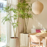 Een lichte, moderne eethoek met een groene potplant van Kunstplant Mahonia (180 cm), een lichte houten tafel en stoelen, beige gordijnen, een hanglamp in de vorm van een papieren lantaarn en natuurlijk zonlicht dat door de hoge ramen naar binnen stroomt.