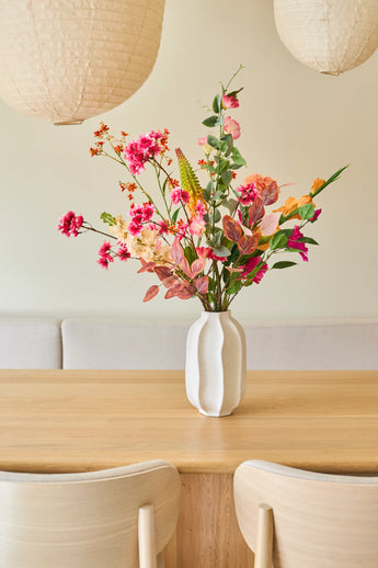 Op een lichte houten tafel staat een witte vaas met een boeket „Zijden kunstbloemen Wild Peach”, samengesteld uit zomerse bloemen. Twee lichte houten stoelen en grote papieren hanglampen maken het minimalistische, gezellige interieur compleet.