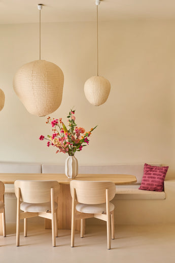 Een minimalistische eethoek met een tafel en stoelen van licht hout, beige muren, drie hanglampen in de vorm van papieren lantaarns, een vaas met het boeket ‘Wild Peach’ van Zijden kunstbloemen dat voor een levendig kleuraccent zorgt, en een ingebouwde bank met daarop een decoratief roze kussen.