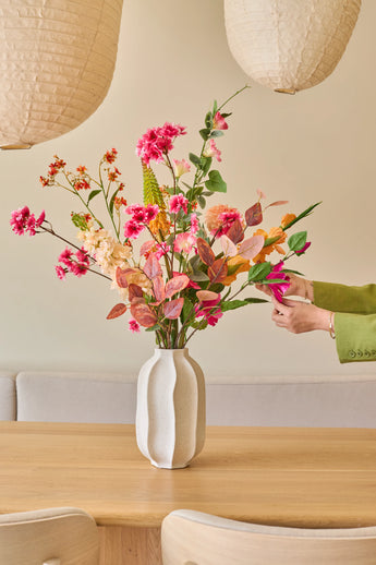 Iemand met een groene mouw schikt het boeket ‘Zijden kunstbloemen Wild Peach’, met kleurrijke zomerse bloemen, in een witte vaas op een lichte houten tafel, met papieren lantaarns en een beige bank op de achtergrond.