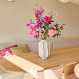 Op een lichte houten tafel in een lichte, moderne eethoek met neutrale tinten en zacht zonlicht dat naar binnen stroomt, staat een witte vaas met het boeket ‘Purple Dawn’ van Zijden kunstbloemen.
