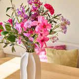 In een witte keramische vaas staat het boeket ‘Purple Dawn’ van Zijden kunstbloemen, een levendig arrangement van roze, paarse en magenta bloemen; de vaas staat op een lichte houten tafel met kleurrijke kussens op de achtergrond.