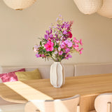 Op een lichte houten eettafel staat een witte vaas met het boeket ‘Purple Dawn’ van Zijden kunstbloemen – een prachtig boeket van zijden bloemen in roze en paarse tinten. Beige stoelen, een ingebouwde bank met kussens en grote papieren lantaarns maken de gezellige, zonovergoten kamer compleet.