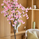 De Kunst Kersenbloesem 180 cm roze, met zijn delicate roze bloesems, staat in een moderne woonkamer met houten planken, keramiek, een beige fauteuil met een groen kussen en een minimalistische inrichting.