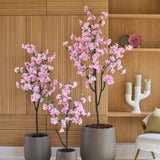 Drie Kunst Kersenbloesem-bomen van 120 cm, versierd met delicate roze bloesems, staan voor een houten lambrisering, naast een beige fauteuil en een moderne witte kandelaar op de plank. Deze elegante opstelling van kunstbloemen geeft elke ruimte blijvende schoonheid en verfijning.