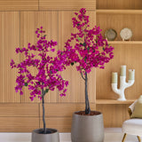 Twee potplanten van het type Kunstplant Bougainvillea van 180 cm, in de kleur paars, met levendige paarse bloemen staan voor een lichte houten wand en planken met decoratieve voorwerpen zoals kaarsen en borden. Rechts staat een beige stoel, die een gezellige sfeer toevoegt aan dit levendige kunstplant-woonkamertje.