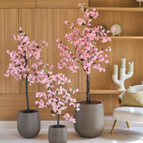Drie potplanten van het type Kunstplant Bougainvillea van 150 cm, in roze met roze bloemen, staan opgesteld voor houten planken, naast een moderne witte stoel met een kussen en een decoratieve kandelaar.