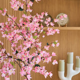 Een kunstplant bougainvillea van 180 cm, roze van kleur met roze bloemen, staat voor houten planken waarop een decoratief bord en een witte kandelaar met drie groene kaarsen staan.