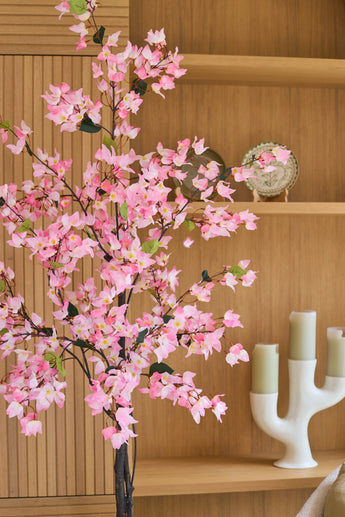 Een kunstplant bougainvillea van 180 cm, roze van kleur met roze bloemen, staat voor houten planken waarop een decoratief bord en een witte kandelaar met drie groene kaarsen staan.