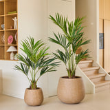 Twee Kunstplant Kentia-palmen van 120 cm, kunstplanten in grote, ronde, beige potten, staan op een lichte vloer bij de trap in een moderne, minimalistische woonruimte met houten planken en een neutrale inrichting.