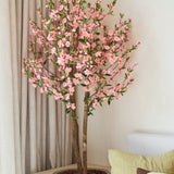 De Kunstplant Kersenbloesem 120 cm roze, met zijn roze kersenbloesembloemen, staat naast een houten tafel in een lichte, moderne kamer, met daarachter een beige gordijn en een lichte bank.