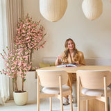 Een vrouw met lang blond haar zit aan een lichte houten tafel, een boek te lezen en glimlachend. Naast haar staat de Kunstplant Kersenbloesem 150 cm roze, waarvan de roze bloesems een levendig accent geven. Hanglampen en een minimalistische inrichting zorgen voor een lichte, gezellige sfeer.