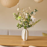Op een houten tafel staat een witte vaas met een elegant boeket kunstbloemen van Zijden, genaamd Claire de Lune. Er staan twee beige stoelen en boven de tafel hangen papieren lantaarns. De achtergrond bestaat uit een zacht crèmekleurige muur met een bank met kussens, wat een minimalistische en gezellige sfeer creëert die doet denken aan de serene sfeer van Claire de Lune.