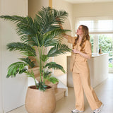 Een vrouw in een beige pak staat binnenshuis, glimlachend, terwijl ze de bladeren van de Kunstplant Phoenix 200 cm aanraakt, vlakbij een trap in een lichte, moderne kamer.