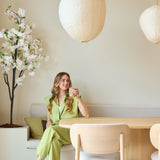 Een vrouw in een lichtgroen pak zit op een bankje aan een houten eettafel, met een kopje in haar hand. Boven haar hangen decoratieve papieren lantaarns en in de hoek staat de Kunst Kersenbloesem 180 cm wit, terwijl er zacht natuurlijk licht door een raam naar binnen valt.