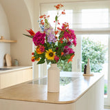 Een witte vaas met het boeket ‘Colourful Spirit’ van Zijden kunstbloemen staat op een licht houten keukeneiland in een lichte, moderne keuken met grote ramen en een neutrale inrichting, en vormt zo een duurzaam en levendig blikpunt.