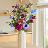 In een hoge, crèmekleurige keramische vaas staat het Zijden kunstbloemenboeket Purple Rain, een arrangement van paarse, blauwe, roze en witte zijden bloemen. Dit handgemaakte boeket staat sierlijk op een licht houten aanrecht, badend in zacht natuurlijk licht – een ware weerspiegeling van Nederlands vakmanschap.