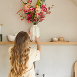 Een vrouw met lang golvend haar zet een vaas met het Zijden kunstbloemenboeket Wild Peach op een houten keukenplank boven een gootsteen. Op de achtergrond is een minimalistische inrichting te zien in neutrale tinten.