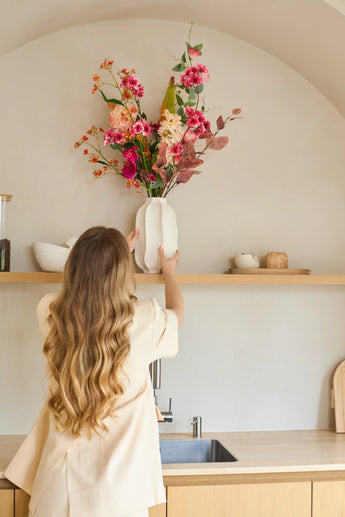 Een vrouw met lang golvend haar zet een vaas met het Zijden kunstbloemenboeket Wild Peach op een houten keukenplank boven een gootsteen. Op de achtergrond is een minimalistische inrichting te zien in neutrale tinten.