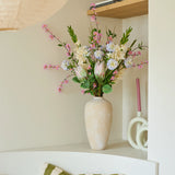 Op een witte plank staat een beige vaas met het kleurrijke boeket kunstbloemen van zijde, ‘Lots of Love XL’, naast roze kaarsen en decoratieve voorwerpen. Op een stoel onder de plank ligt een groen-wit gestreept kussen.
