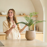 Een vrouw met lang blond haar glimlacht terwijl ze een Kunstpalm Chamaedorea van 48 cm in een pot verzorgt in een moderne, in neutrale tinten gehouden keuken met een minimalistische inrichting.