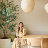 Een vrouw in een beige pak zit glimlachend aan een houten tafel in een moderne, minimalistische kamer met lichte muren, papieren hanglampen die aan het plafond hangen en een Kunstplant Eucalyptus van 180 cm die de ruimte wat groen geeft.