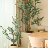 Twee Kunstplant Eucalyptus kunstplanten van 150 cm, geplaatst naast transparante gordijnen en beige muren, gecombineerd met een eettafel van licht hout en stoelen in een minimalistisch, modern interieur.