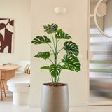 De Kunstplant Monstera van 120 cm staat in een moderne, minimalistische woonruimte met neutrale muren, een ronde spiegel, houten meubels en abstracte wandkunst.