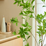 Een kunstplant Pieris Japonica van 150 cm staat bij een raam naast een licht houten kastje, waarop keramische flesjes en een pepermolen staan, wat een minimalistische toets geeft aan de lichte, luchtige kamer.
