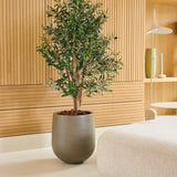 Een Kunst Olijfplant 120 cm UV staat op een lichte vloer naast een beige bank in een moderne kamer met houten lambrisering en minimalistische planken waarop vazen en servies staan uitgestald. Dit is een onderhoudsvrije olijfboom die perfect past bij een stijlvolle, onderhoudsvriendelijke inrichting.