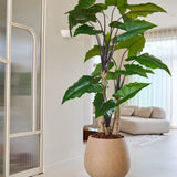 De Kunstplant Alocasia van 240 cm, met zijn brede groene bladeren, staat prominent in een lichte, moderne woonkamer met een lichtgekleurde bank, doorschijnende gordijnen en een minimalistische inrichting.