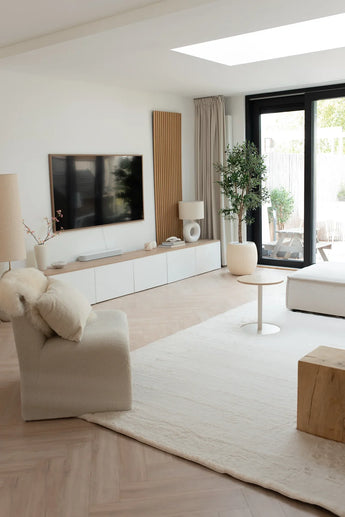 Een moderne woonkamer met neutrale tinten, met een fauteuil met structuur, een lage witte bank, een houten salontafel, een groot vloerkleed, een tv aan de muur, grote ramen voor natuurlijk licht en een Kunst olijfboom UV 160 cm als potplant.