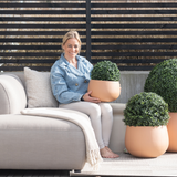 Een vrouw zit op blote voeten op een lichtgekleurde buitenbank, glimlacht en houdt een Kunstplant Buxus romeinse stijl UV D30cm vast. Twee soortgelijke kunstplanten staan vlakbij, met houten lattenhekwerk en terrasdecor op de achtergrond.
