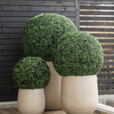Drie Kunstplant Buxus romeinse stijl UV D65cm plantenbakken met netjes gesnoeide groene struiken staan op een houten terras naast een modern donkerhouten privacyscherm.